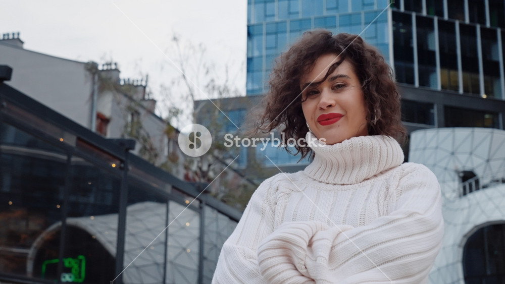 Portrait gorgeous young woman with curly hair standing in modern city street looks at camera smiles. Charming girl wearing white pullover autumn outfit enjoys life posing backdrop downtown skyscrapers