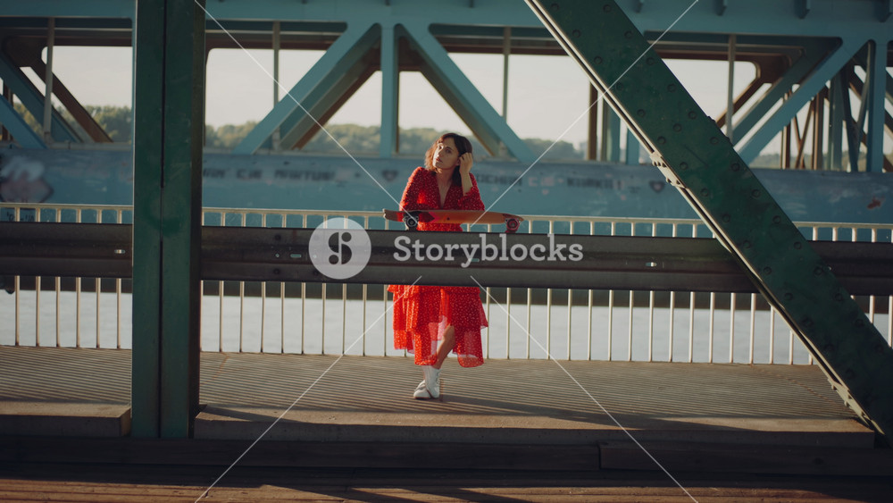 Relaxed brunette woman in red dress posing with skateboard leaning on bridge with massive green girders enjoying summer evening, stylish caucasian female skateboarder, industrial urban background