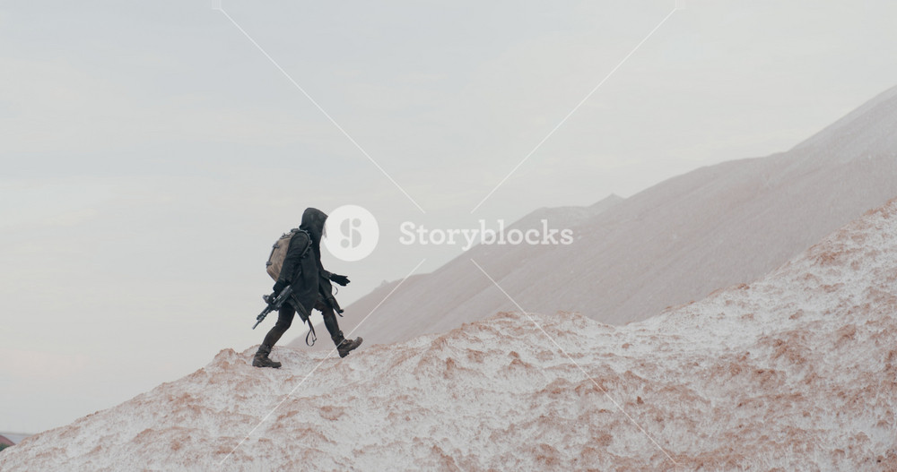 Wide shot post apocalyptic woman in grunge clothes hooded black mantle protective gas mask respiratory walking up hill in white desert. Cyberpunk soldier holding assault gun. Future female survivor