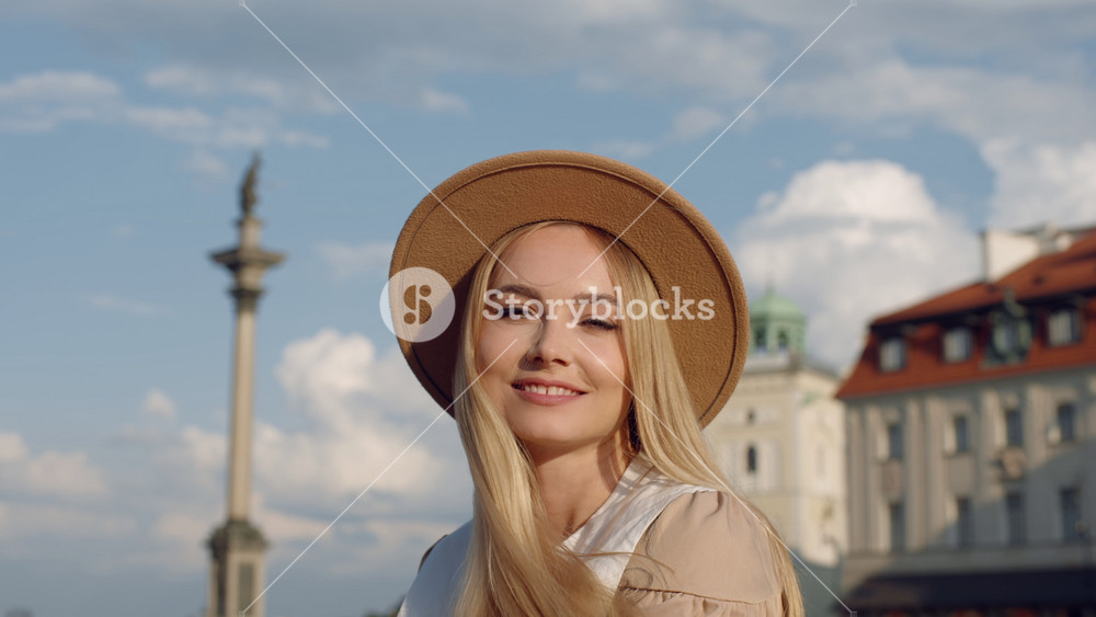 Portrait beautiful young woman posing while standing in old town square. Cute female wearing fedora hat and dress looking at camera and at cityscape, sunny summer evening cloudy blue sky on background