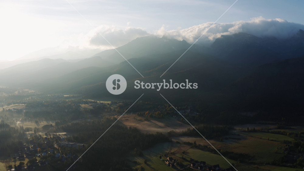 Aerial foggy autumn morning in alpine village lit with bright sunlight. Drone shot epic mountain peaks covered with rolling clouds in sunrise light. Incredible landscape beautiful valley from above