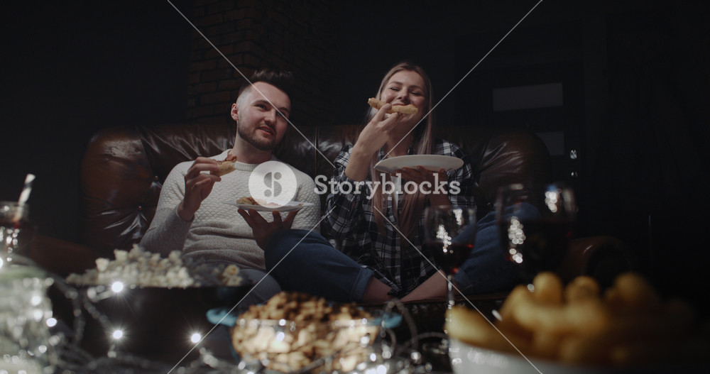 Happy millennial couple sitting on couch having fun watching TV show or movie at home in evening. Handsome man and attractive woman eating pizza, talking and smiling each other. Wide angle dolly shot