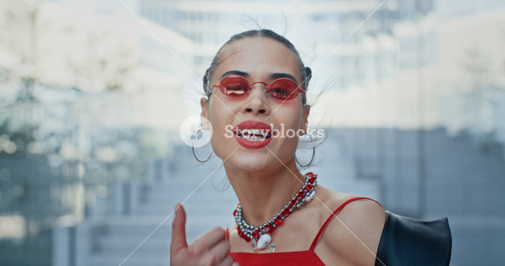 Portrait of stylish gorgeous cheeky woman, seductive happy female wearing cool red eyeglasses smiles to camera and flirts standing in modern business center overlooking high rise glass skyscrapers