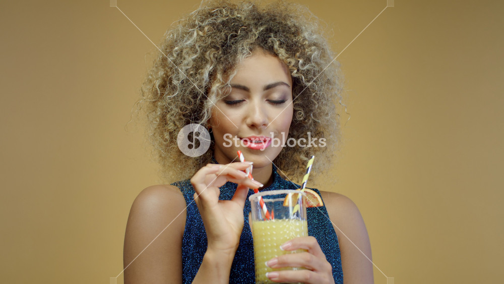 Portrait beautiful mixed race woman in blue shiny blouse drinks orange cocktail through paper straw on yellow background. Joyful african female enjoys holding glass of refreshing drink and smiling