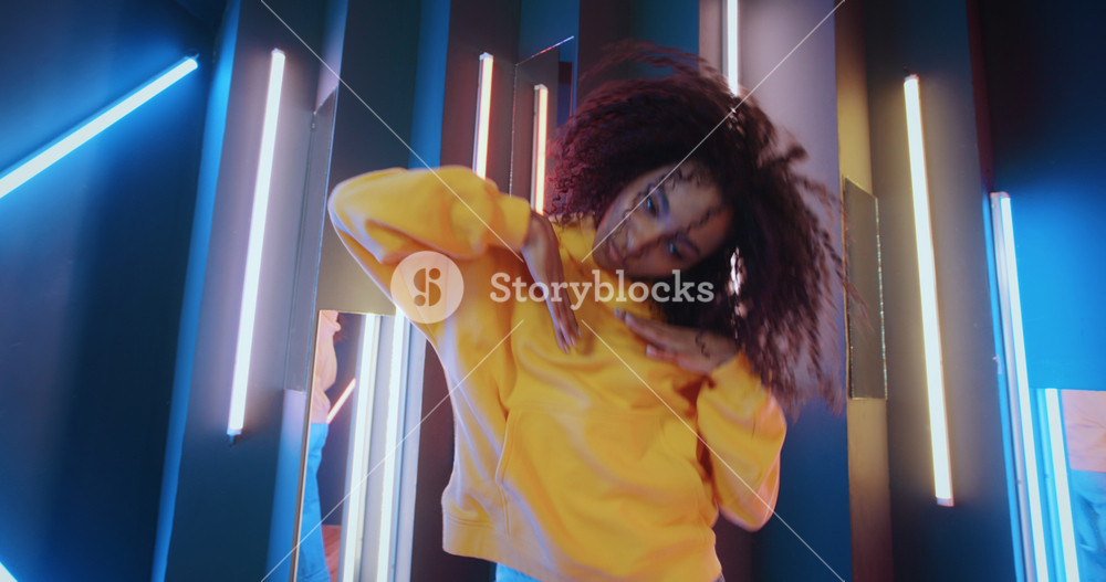 Active urban african american black woman in yellow hoodie dancing by neon lights background in disco club. Cool talented mixed race female dancer feeling free moves rhythmically to hip-hop music beat
