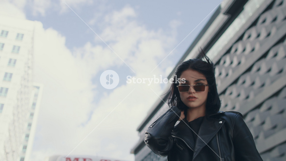 Confident young brunette woman in trendy stylish look wearing black leather jacket and sunglasses looking at camera standing by building in city street, touches her hair. Lifestyle, fashion, urban