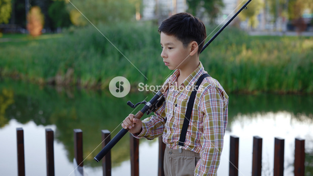 Asian boy goes fishing on the pier, dressed in special fishing clothes, holding a fishing rod in his hands. Side view of the asian boy goes