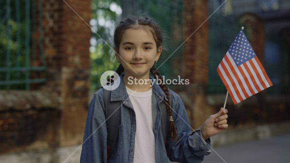 A little girl standing outside near school with american flag. Patriotic female child holding in hands a flag. Patriotic concept