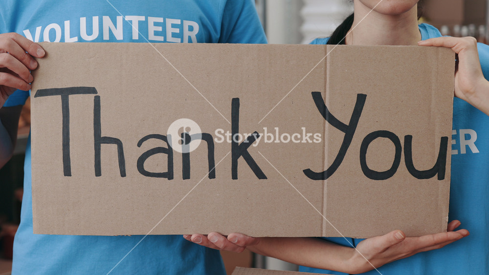 Close up of caucasian volunteers holding card with lettering thank you while standing at food bank warehouse. Young man and woman