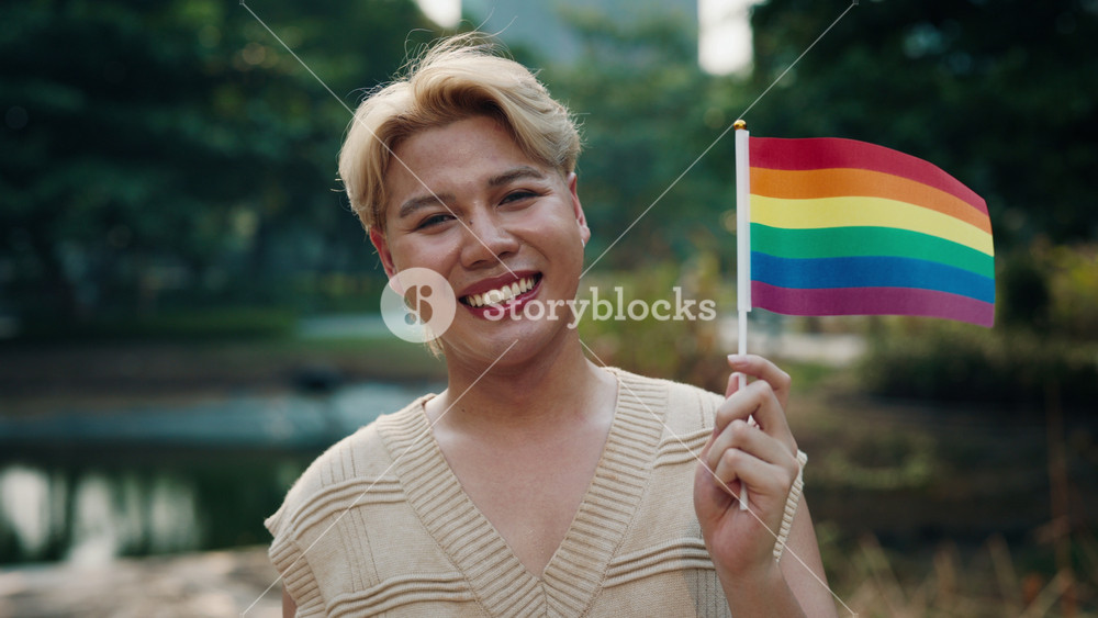 Joyful Asian man gay with dyed blond hair smiles holding rainbow flag in city park. Young Japanese male proud to show as LGBT community