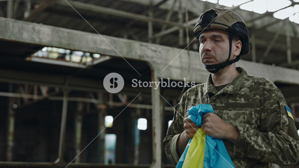 Tired emotional soldier in camouflage uniform and cap holding Ukrainian flag with love and praying. Blur background of destroyed factory