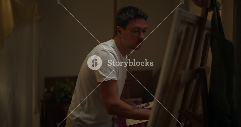 Male artist takes thick paint from his wooden palette with a small brush and applies it in large strokes to a canvas during the creative