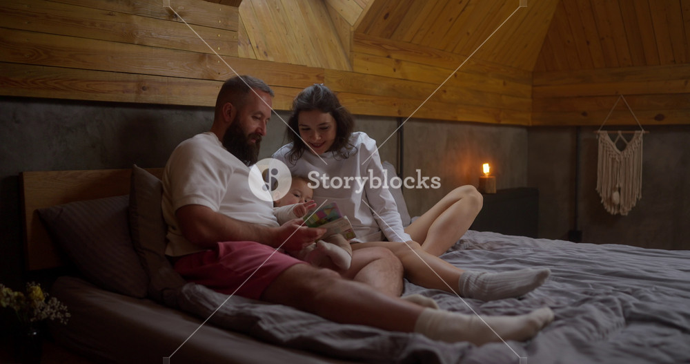 Happy brunette woman lies on the bed with her husband and reads a book to her little daughter in a cozy wooden room