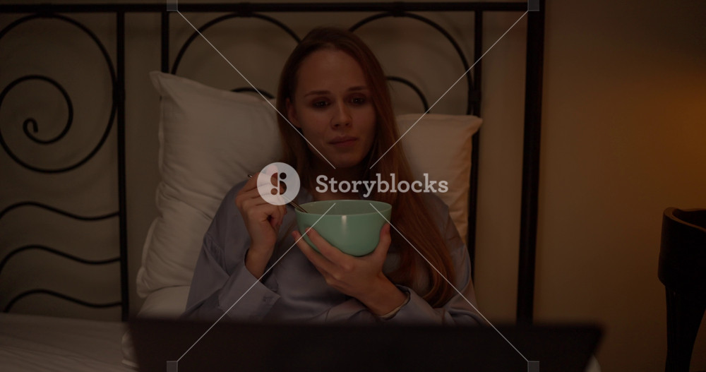 A blonde girl in pajamas lies on the bed and watches a movie on a laptop during her dinner before going to bed at night