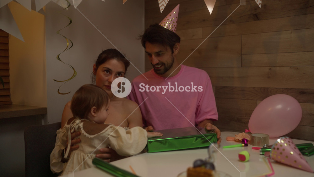 Happy brunette man in a pink t-shirt with his wife and little daughter inspecting a gift in a green glossy package during a birthday party