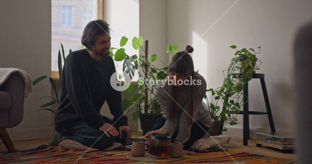 Girl pouring hot tea from teapot and giving mug to her boyfriend during cozy tea party on floor in cozy apartment at home. Cozy relaxation