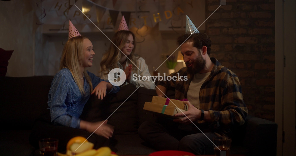 Two happy girls friends in festive caps give gifts to their friend the birthday boy during a party at home in the evening