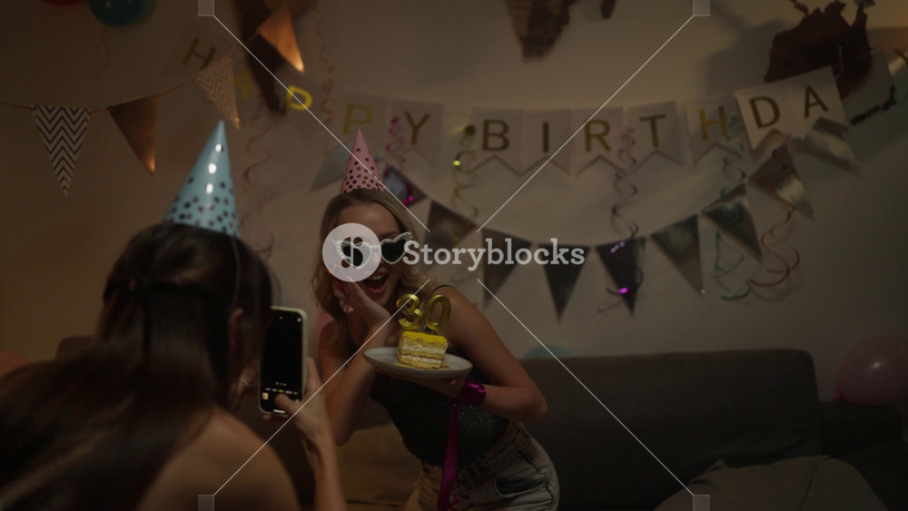 Over shoulder happy blonde girl in pink party cap posing and smiling holding cake with festive candles during birthday party in the evening