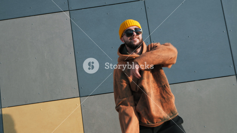 Dynamic young man dancer in streetwear performs hip-hop dance move in front of modern, tiled wall with geometric patterns in sunny day autumn. Passion energy, urban culture and street art style