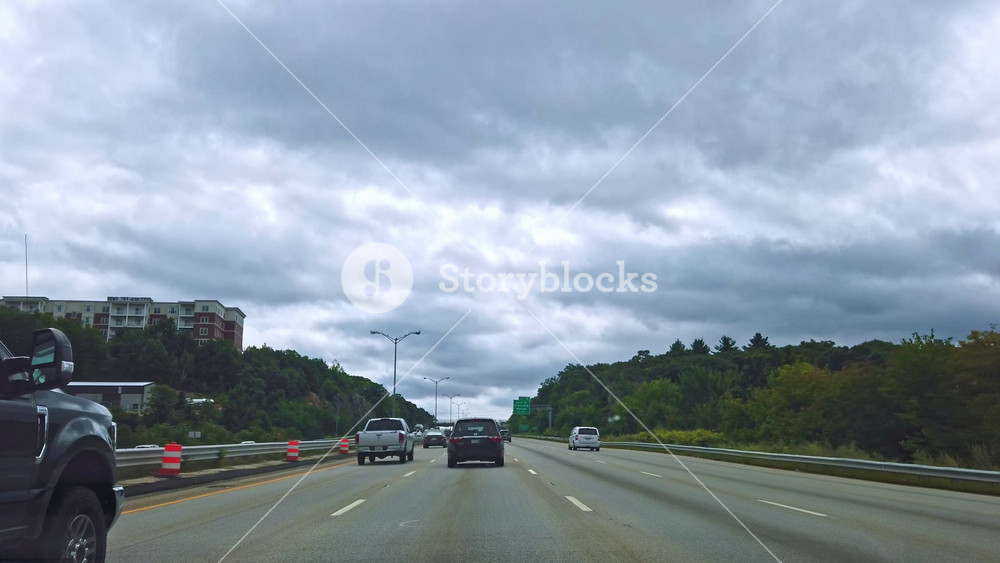 BOSTON, MASSACHUSETTS - SEPTEMBER 11, 2019: Cars drive on a route near the city. Rainy clouds with purple sky. Green forest and stone. Car view