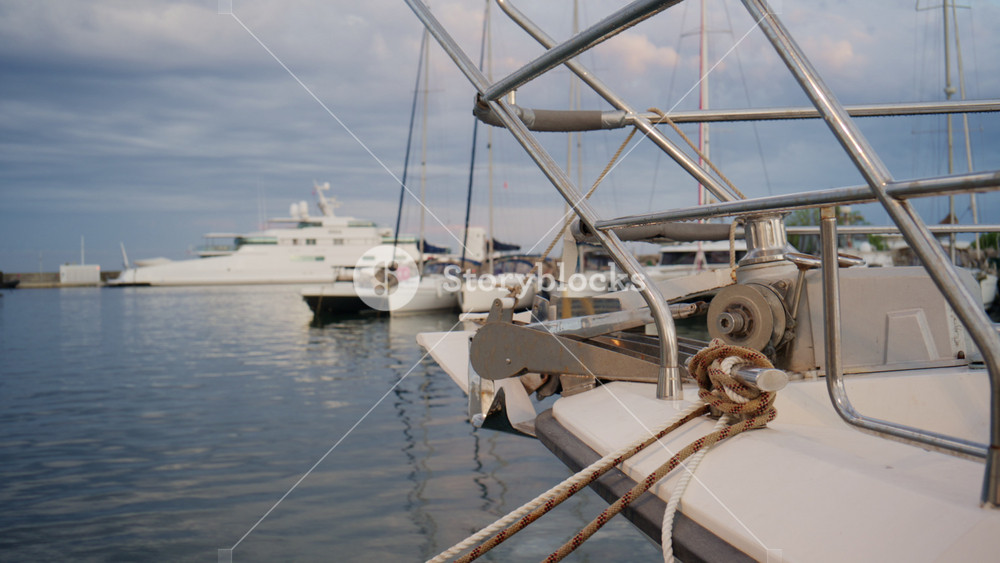 Close up of a sailboat bow, anchor, ropes, and metal railings at Port Vauban marina in Antibes, with yachts and calm Mediterranean water in the background