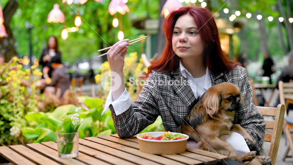 View of a young woman eating poke bowl with vegetables and fruits using chopsticks while holding her dog on the knees. Park with a lot of greenery and illumination. Slow motion