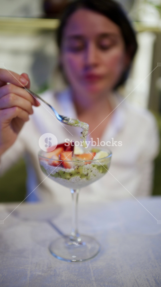 A woman holds a spoonful of chia pudding in front of a dessert glass filled with white chia seed pudding, green kiwi puree, sliced strawberries, and banana. Vertical