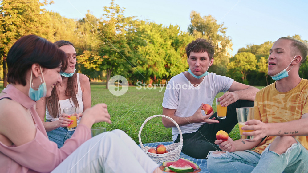 Group of friends in protective medical masks having fun at a picnic blanket with goodies in a park during the pandemic. Corona Virus idea