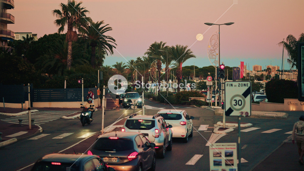 Cannes, France - November 3, 2025: Busy coastal boulevard lined with palm trees at dusk, with cars, scooter and pedestrians moving through traffic under a pastel sky and rising moon