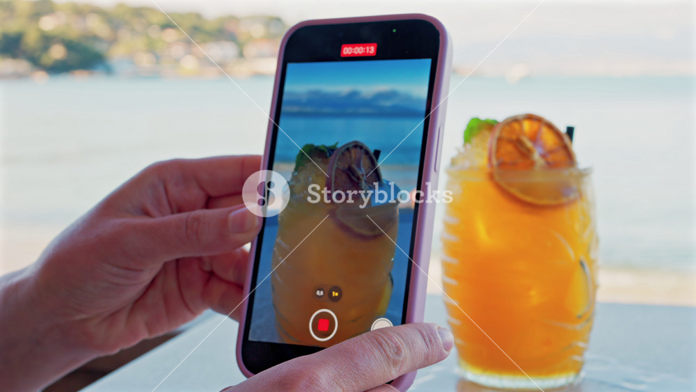 Close up of a woman's hand filming an orange cocktail on her phone with a blurred view of the sea on the background