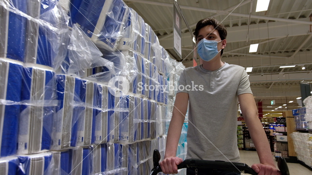 CHISINAU, MOLDOVA - JUNE 5, 2020: Young man with a protective medical mask walking in a supermarket near the shelves with toilet paper and looking around. Corona Virus idea