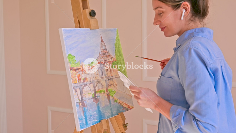 Caucasian woman painting a picture with a palette in her hands and listening music with earphones indoors. Slow motion