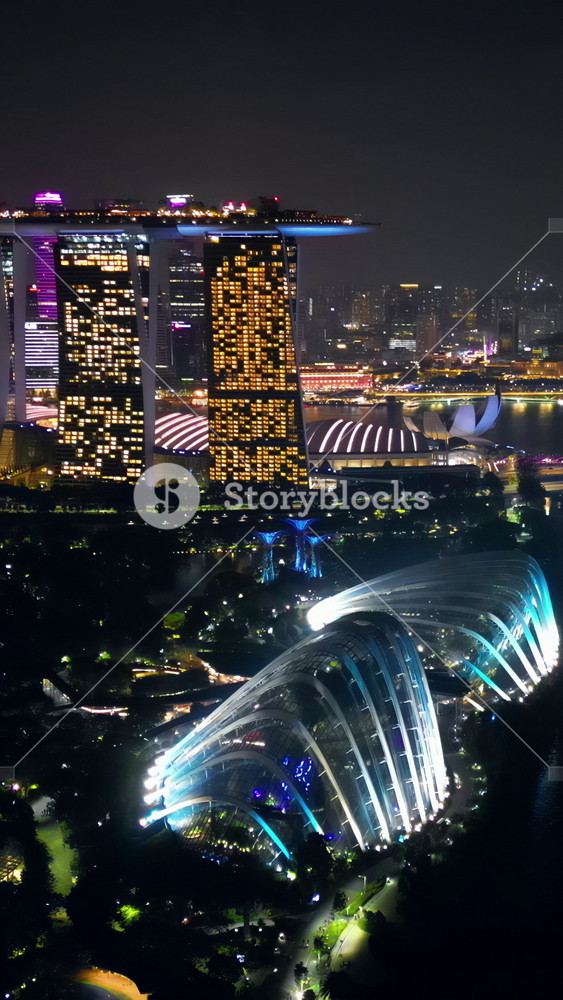 Aerial drone view of Marina Bay in Singapore at night showing Marina Bay Sands, the Singapore Flyer observation wheel, and illuminated waterfront skyline. Vertical