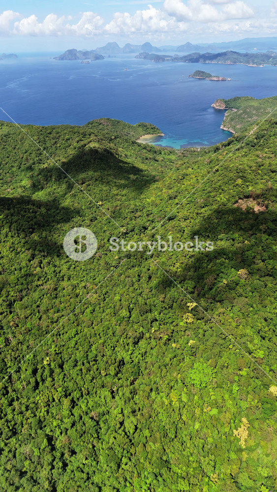 Vertical aerial drone view of dense jungle-covered hills in the foreground with blue sea and distant island silhouettes beyond, Catpo Beach, Liminangcong, Taytay, Palawan, Philippines.