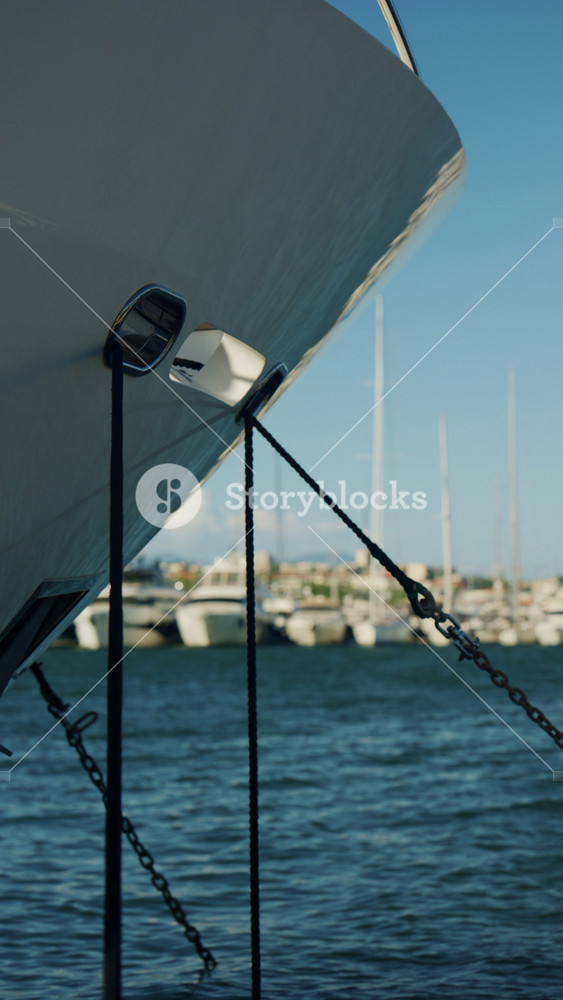 Close up of a large yacht bow with mooring ropes and chains, with sailboats and marina buildings blurred in the background