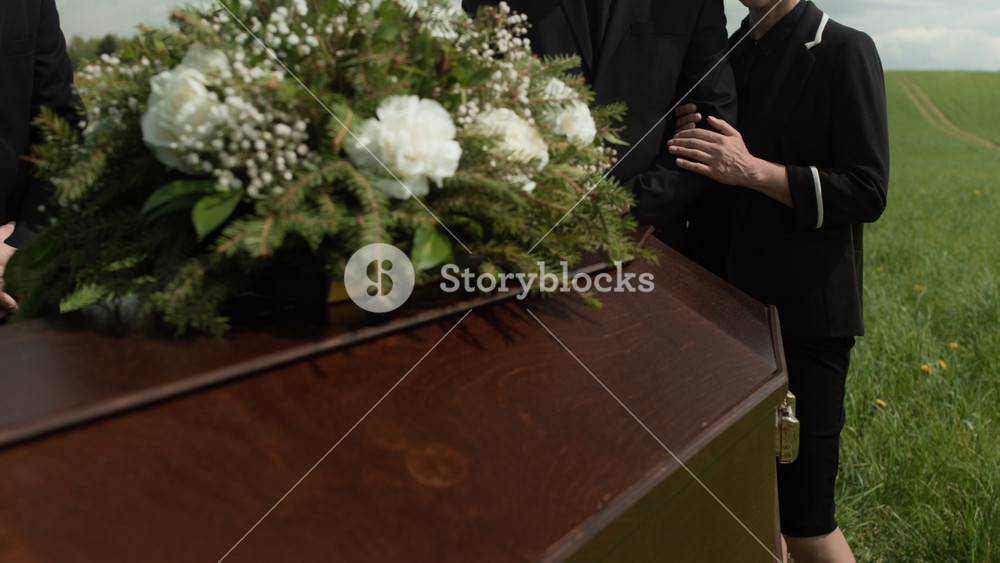 Midsection of diverse family members or friends wearing black clothes standing near coffin decorated with flowers at outdoor funeral supporting unrecognizable Black woman holding folded American flag