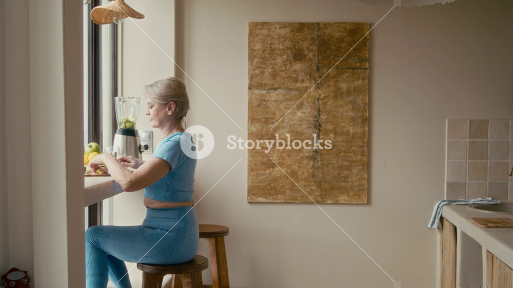 Side view waist up of serene mature Caucasian woman in blue activewear sitting by window, adding fresh green fruits and lemons to her blender while enjoying sunlight in her bright, cozy kitchen