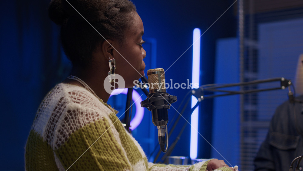 Young Black female radio host listening carefully to her young Caucasian male guest speaking in microphone sitting at table in professional recording studio with ambient blue backlight