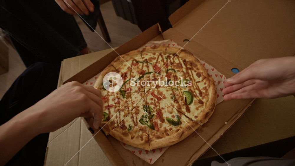 Dolly out shot of four diverse friends relaxing on floor and toasting with takeaway pizza slices, sitting among cardboard boxes in bright apartment on moving day