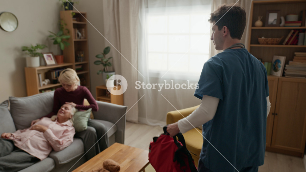 Young adult Caucasian male nurse opening medical bag to treat sick senior woman lying on sofa with middle aged daughter. Ideal for home healthcare services and emergency response advertising