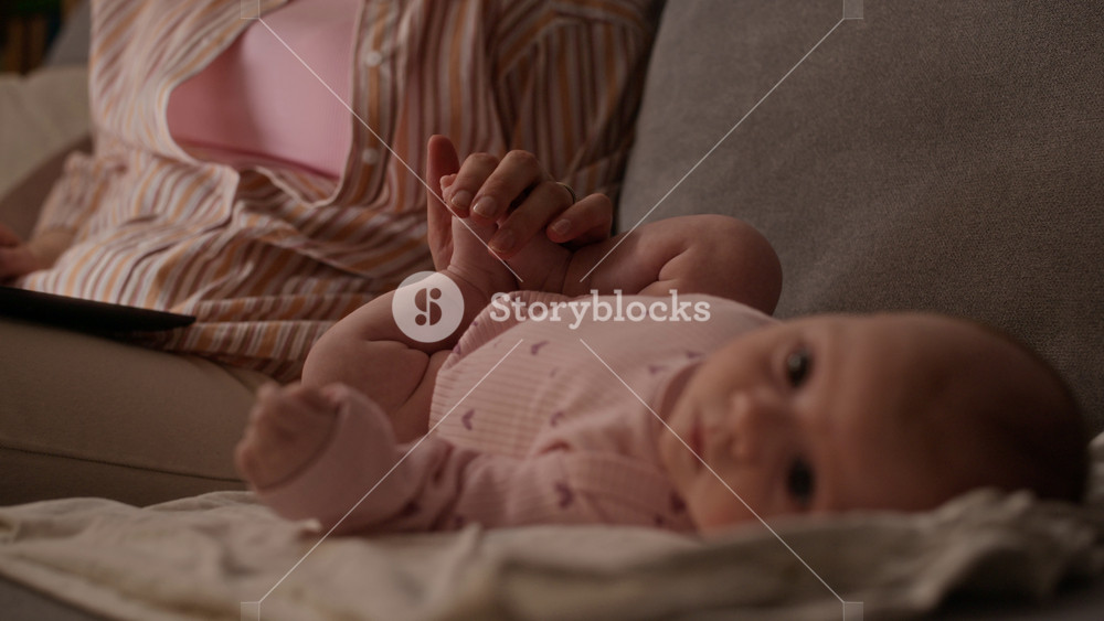 Rack focus shot of infant Caucasian girl wearing bodysuit lying on couch near unrecognizable mother working on laptop from home at night