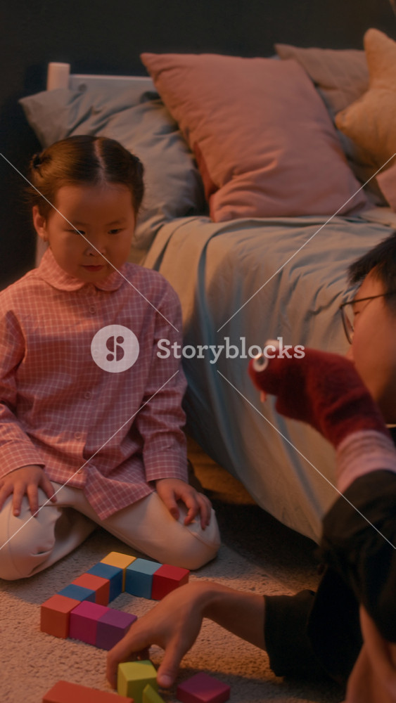 Vertical shot of Asian father and his pretty little daughter sitting on rug near cozy child bed and playing with sock puppet during evening playtime in cozy home setting