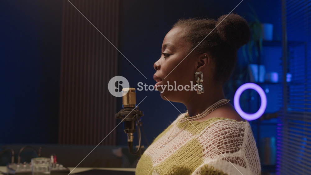 Side view chest up shot of young confident African American woman with shiny earrings speaking in mic during podcast sitting in bright modern recording studio with glass partition and blue backlight