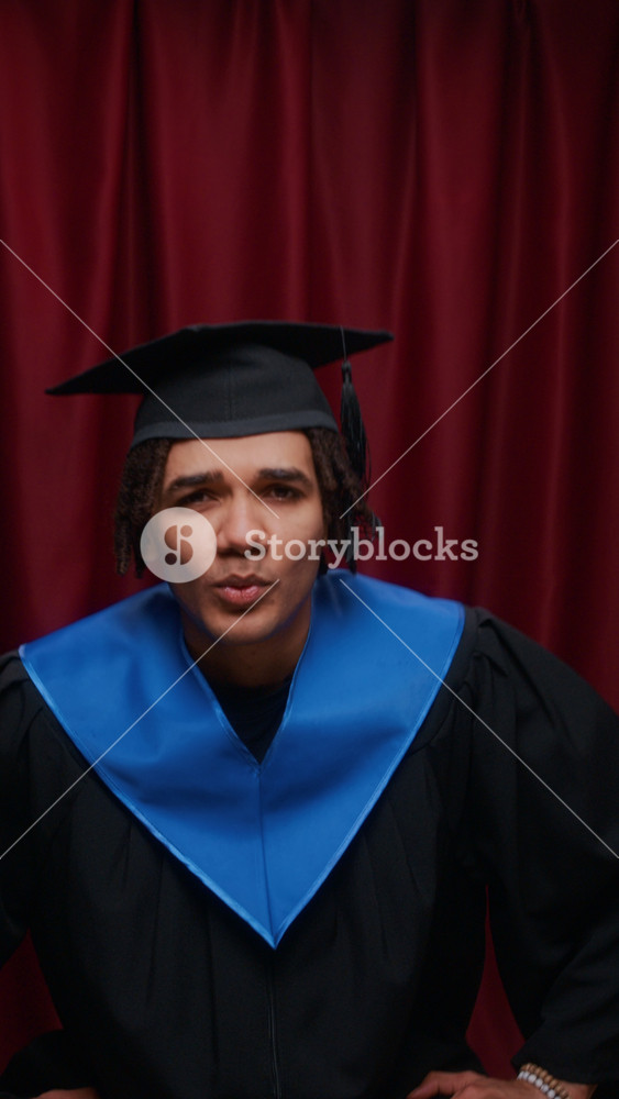 Vertical waist up of proud young Biracial male graduate adjusting his cap while posing in photobooth, wearing black graduation gown with blue stole, set against red curtain backdrop