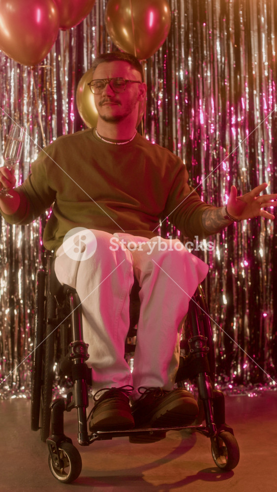 Vertical full portrait of cheerful young Caucasian man with disability looking at camera while dancing, then raising champagne glass at birthday party with balloons in background