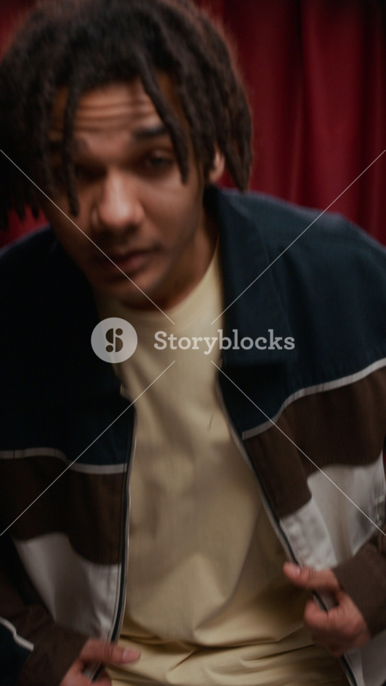 Vertical waist up of young Biracial man with short locs in color block jacket making playful shaka hand signs and puckering his lips in front of deep red curtains inside photo booth