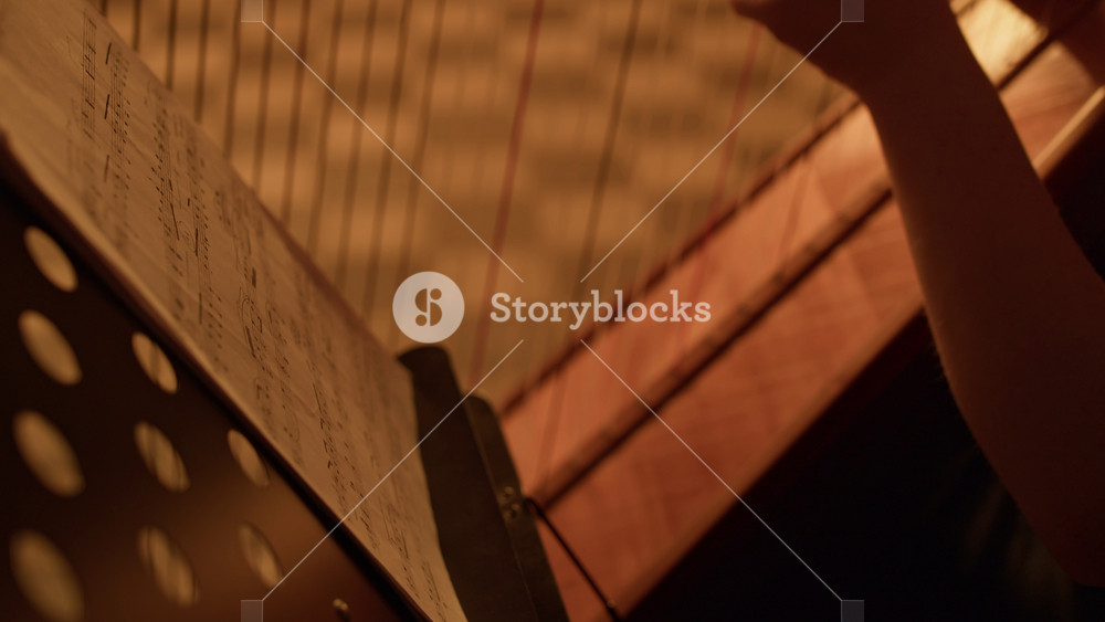 Side view cropped shot of young female harpist sitting near harp and turning pages of sheet music on music stand while playing harp in dim lit room