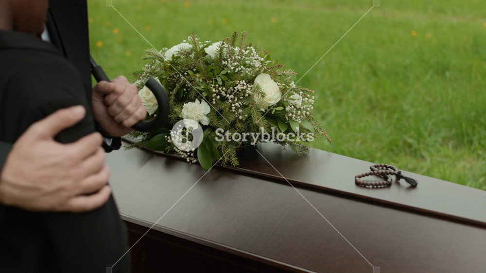 Cropped slowmo of unrecognizable man holding umbrella hugging grieving Black woman standing near closed wooden casket decorated with flowers at outdoor funeral