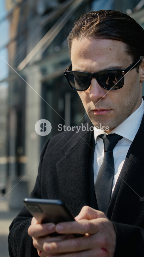 Vertical shot of young adult Caucasian businessman in sunglasses standing outdoors near office building, typing on smartphone. Clip is ideal for corporate communications or mobile banking ads