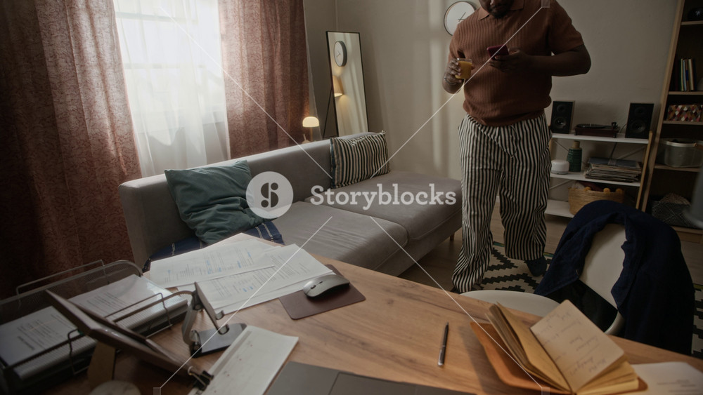 Handheld shot of young African American man walking with juice, sitting at desk, organizing papers, writing in notebook. Clip is ideal for remote work, freelance productivity, home office lifestyle
